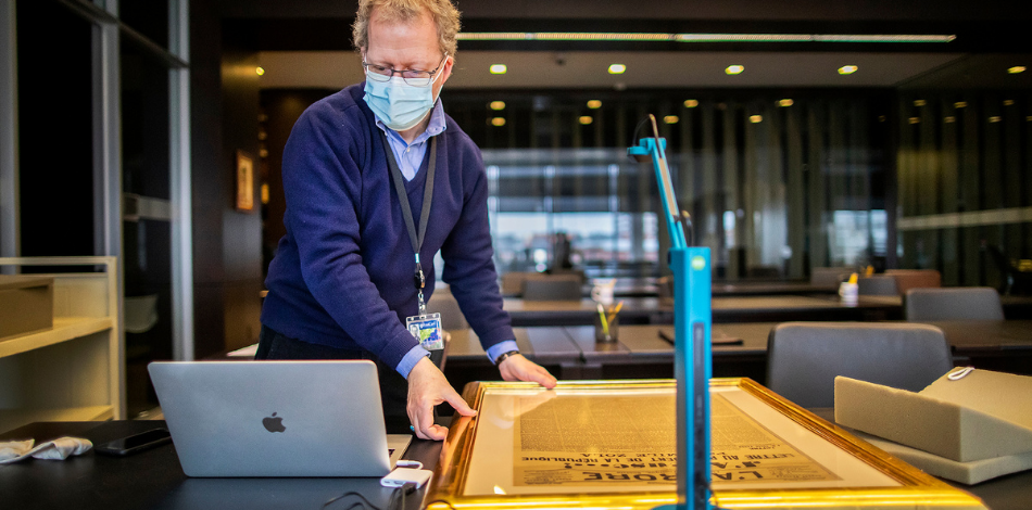 A man wearing a surgical mask looks down at a computer. Beside him is a large historical broadside with a bright blue document camera pointing at it.
