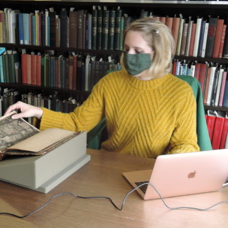 A person wearing a face mask turns the page in a manuscript while streaming on a laptop.