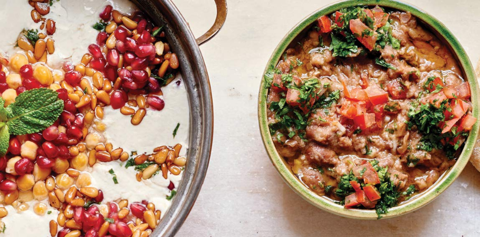 Two bowls of food: the one on the right is a bubbling curry-like dish topped with brightly-colored tomato and parsley; the one on the left is a creamy white soup topped with chickpeas, pomegranate, and pine nuts  