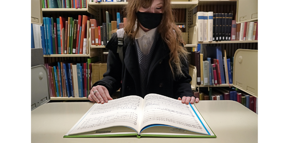 A woman wearing a face mask stands in front of an empty bookshelf, looking down at a large book that is resting open on the shelf