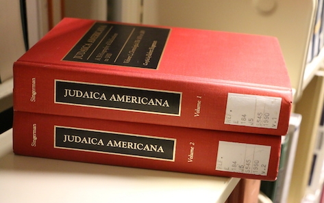 A photo of volumes 1 and 2 of Robert Singerman's JUDAICA AMERICANA stacked on a library shelf. The books have bright red ocvers and the title written in gold inside a black box.