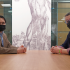 Manuel and Richard sit at a table, wearing masks and looking at the camera. A medical illustration decorates the wall behind them