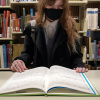 A woman wearing a face mask stands in front of an empty bookshelf, looking down at a large book that is resting open on the shelf